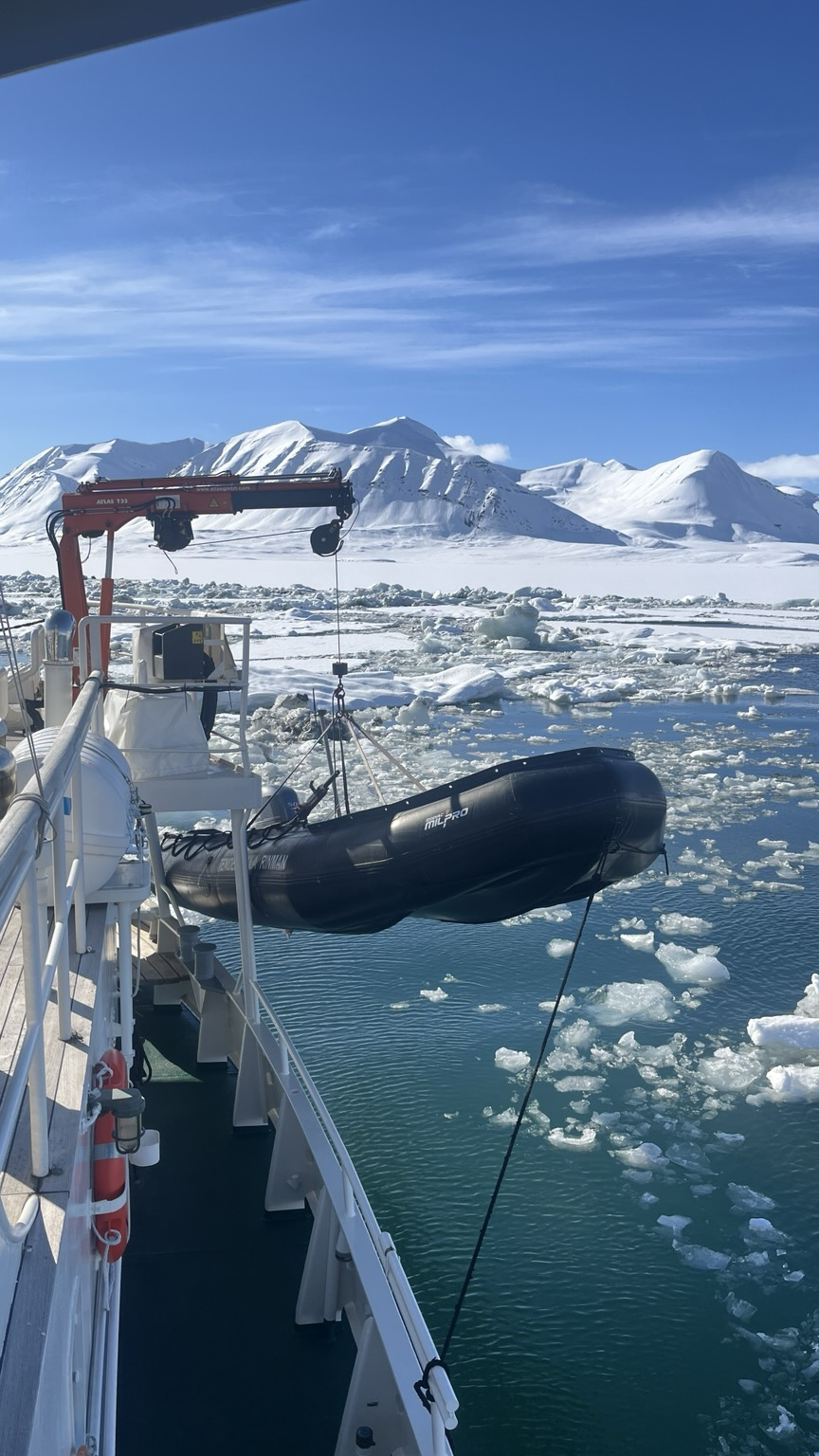 A black inflatable boat hanging in a crane over broken up sea ice. Snowy mountains in teh background.